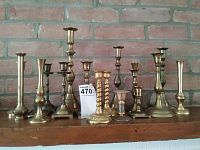 Wide shot showing 13 assorted brass candle sticks in various styles and sizes arranged on a wooden surface in front of a brick wall.