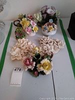Overview of all ceramic floral candle holders and arrangements including a pair of ruffled candle holders, a pitcher-style candleholder, and several small floral pieces arranged on a white surface.