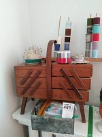 Front view of vintage wooden sewing box with handle, pincushion with pins on top, and several stacks of thread spools on wooden rods next to it. Box of buttons partly opened below the sewing box.