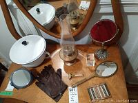 Photo showing brass oil lamp with glass chimney, vintage standing vanity mirror, standalone magnifying mirror on ornate base, vintage hand-held mirror, brown leather gloves, vintage shaving razor, and knitting guide on wood surface