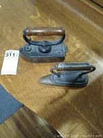 Pair of vintage sad irons on wood surface; one slipper iron with pointed nose and wooden handle, one cast iron reservoir style with handle attached.