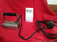 Small antique Baby Betsy Ross electric iron with black handle and knob and tube fuse plug shown from a side angle on a red background with lot number card.