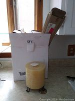 Large yellow pillar candle on a small black metal holder sitting on a kitchen counter with a white box behind it.