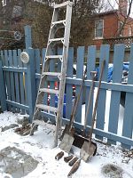 Wide shot of 7-foot aluminum step ladder with three shovels and some wedges on snowy ground by blue fence.