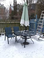 Photo showing outdoor round metal table with green and white umbrella, snow on table surface and ground.