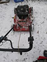 Top front view of the red Troy-Bilt lawn mower on snow-covered ground, showing engine and handlebar with pull start cable untied and throttle cable.