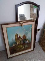 Antique wall mirror with dark wood frame shown leaning against wall next to framed landscape painting.