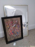Frosted mirror with metal frame standing behind a framed Mexican folk art bark painting featuring colorful birds and flowers.