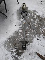 Photo of the full length of the gas trimmer showing shaft, handle, and cutting head lying on snow-covered ground.