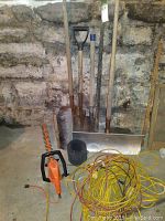 View of all items against a stone wall: two yellow extension cords, scraper shovel, edge shovel, tiller tool, broom head, black mesh roll, and orange electric hedge trimmer.