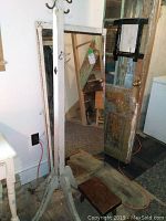 Wooden coat tree standing beside a rustic door and a mirror leaning against the wall, showing the coat tree with four feet needing regluing and top sawed off.