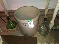 Wide shot showing large beige crock with '6 Imperial' marking, flanked by two clear glass jugs, one with a woven basket.