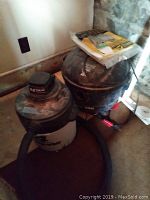 Two shop vacuums placed on the floor against a wall, one white and black with hose attached, the other black with papers on top.