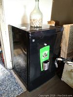 Black GE bar fridge front view showing green mounted cap remover and cap catcher basket. A large glass bottle filled with corks on top of the fridge.