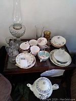 Full lot on wooden table showing cups, saucers, glasses, teapot, and vintage oil lamp.