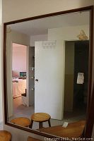 Large rectangular mirror with teak wood frame hanging on a wall, reflecting room interior with stools.