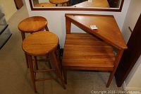 Photo showing two round wooden stools and one teak side table with triangular top shelf and square bottom shelf under a mirror.