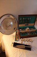 Photo showing the large silver plate platter, wooden case of Community Stainless flatware set, hallmarked sterling handle cake server and bottle opener, with some spoons laid out.
