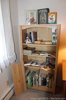 Front view of light wood bookcase with shelves holding books, magazines, puzzles, and cat figurine, with a detached wooden shelf panel leaning beside it.