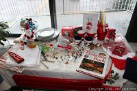 Photo of assorted Christmas holiday tableware and decorations arranged on a table including plates, festive mugs, wine charms, holiday linens, a Christmas book, and a lit basket