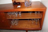Photo showing various old fashioned glasses, wine glasses, and other glassware stored inside a wooden cabinet shelf with a mid-century design.