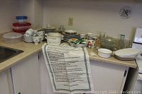 Wide view of kitchen counter with various dishes, glassware, and a printed paper listing contents in foreground