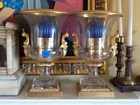 Two clear glass candle holders with brushed gold trim and gold decorative side handles, each containing a blue candle. Positioned on a shelf with other candle holders nearby.