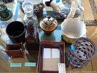 Photo of 9 items on a glass surface including vases, canister, decanter, statue liquor dispenser, picture frames, and a small container