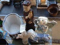 Five items on a glass table include a white ceramic planter with stand, tall wooden statue, cherub statue, reclining baby statue, and coral piece