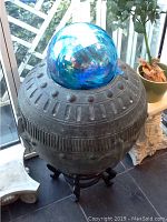 Metal art round vessel topped with blue glass globe on dark wooden stand
