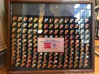 Full view of wood framed 1984 Official Olympic Coca Cola International Flag Pin Series with multiple pins arranged in rows on black backing.