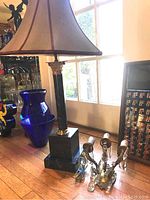 Full view of marble lamp with heavy square base and tall column, topped by large gray and burgundy fabric shade alongside brass three-light wall sconce with hanging crystals on floor