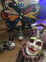 Butterfly-shaped painted case behind three Mardi Gras jester masks on wooden floor
