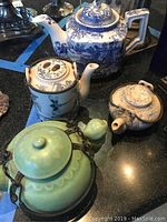 Four ceramic teapots arranged on a black surface, showing different styles and colors including blue and white floral, cream with brown flowers, green ceramic with metal handle, and beige patterned.