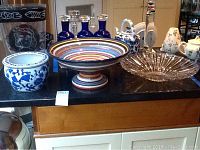 View of three items on black surface including clear glass beveled bowl, multicolored pedestal bowl, and blue and white planter with floral design, along with other background objects.