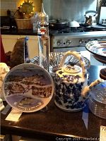 Three items on a black surface: a glass thermometer with floating gauges, a souvenir Kusadasi plate, and a blue and white floral teapot with bamboo handle