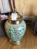 Green ceramic vase with pink and red flowers, brass top and bottom, shown from front angle on wooden floor near furniture