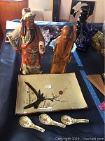 Photo showing two Asian figurines (ceramic and wooden), rectangular dish with bird and floral design, and three floral pattern ceramic spoons arranged on a table.