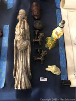Overview of Asian collectible lot showing large soapstone man figure, brass candle holders, porcelain figurine, wooden carved head, trinket box, and carved pig.