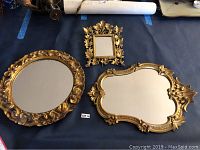 All three mirrors laid out on a blue surface: one round gold gilt wood framed, one rectangular gold metal framed with stand, one irregular shaped gold gilt wood framed.
