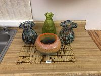 Four vintage art glass pieces on counter: two ruffled multicolored vases, one green bottle-shaped vase, one round gradient bowl.