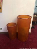 Pair of large empty amber translucent cylinder floor lamps shown side by side on burgundy carpet against white wall, unlit.