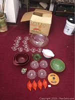 Full lot shown with punched glass bowl, glasses, ladle, and various ceramic and glass serving dishes spread on red carpet with box in background.