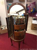 Wooden jewelry box standing on carpet, with mirror, three drawers and side door open showing jewelry inside and draped over top and door.