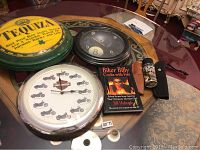 Photo showing all items in the lot arranged on a glass table: two advertising clocks, one wall clock, cookbook, spice bottle, and bicycle model.