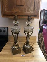 Photo showing front view of both antique crystal lamps on wooden countertop with bronze bases and crystal reservoirs and finials.