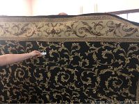 Full view of the large area rug with black background and beige scroll pattern held upright for display.