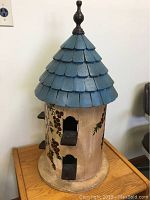 Front view of bird house showing two entry holes with perches, hand painted floral grapevine design, blue shingled roof with finial, placed on wooden surface against plain wall.