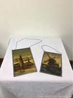 Both vintage reverse painted glass artworks lying flat on white fabric, showing their metal frames and hanging chains.
