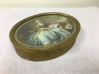 Photo showing oval brass box with detailed embossed designs and glass lid featuring painting of Victorian woman.